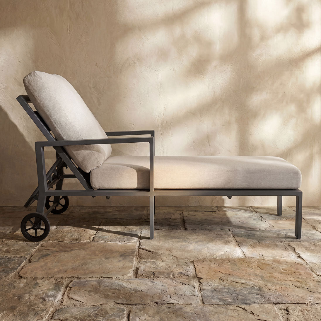 Gray metal outdoor chaise lounge with beige cushions and wheels on a stone patio by Paddy O' Furniture.