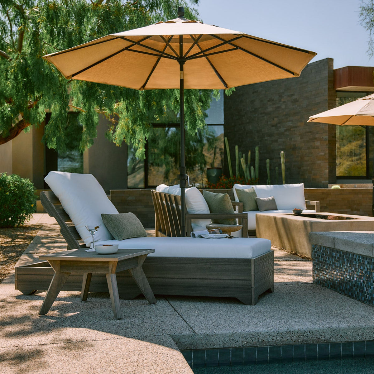 Modern patio lounge set with grey wood chaise, white cushions, tan umbrella, and side table by a pool.