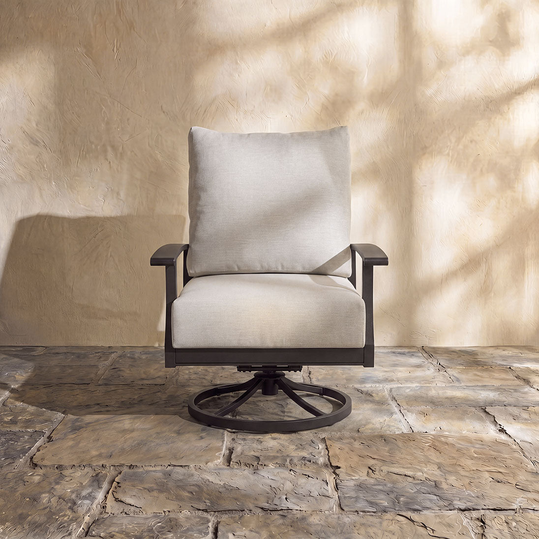 High-back outdoor swivel rocker with plush off-white cushions and a durable dark metal frame on a stone patio.